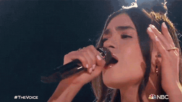 a close up of a woman singing into a microphone with the nbc logo behind her