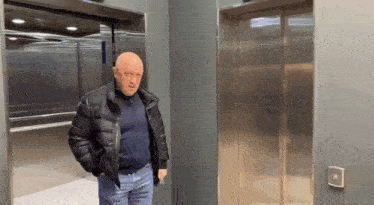a bald man in a black jacket is standing next to an elevator .