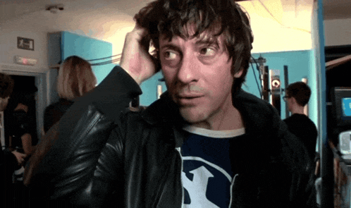 a man scratches his head while wearing a black jacket and a blue shirt with an arrow on it