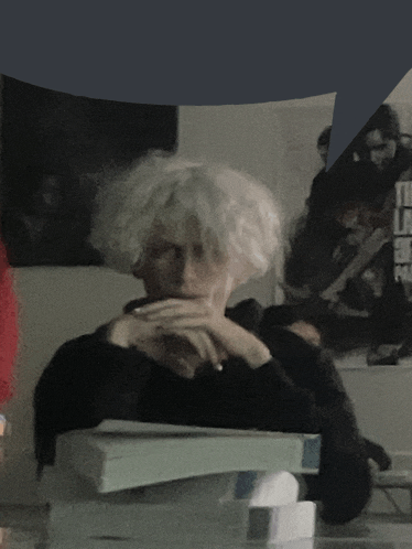 a woman with white hair sits at a desk with her hands on her chin
