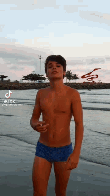a young man without a shirt is standing on a beach wearing blue swim trunks .