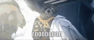 a man wearing a white jacket and gold chains is standing in a dark room .