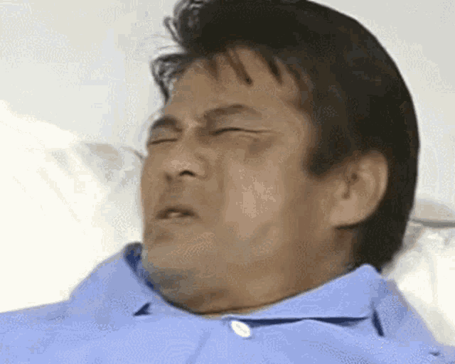 a man in a blue shirt is laying in a hospital bed with his eyes closed and making a funny face .