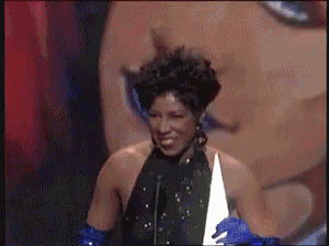 a woman in a black dress and blue gloves is smiling while speaking into a microphone