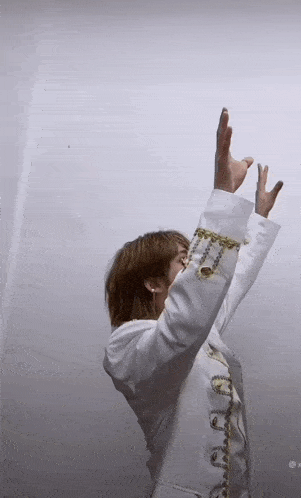 a woman in a white jacket with gold chains on the sleeves holds her arms up in the air