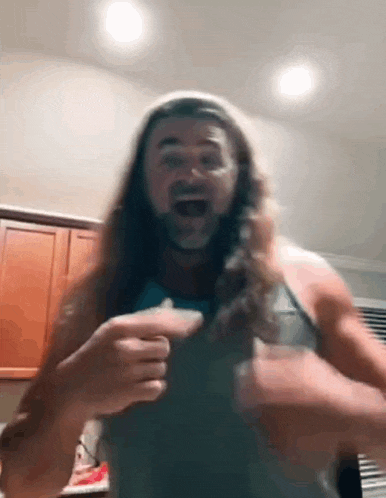 a man with long hair and a beard is standing in a kitchen holding a remote control .