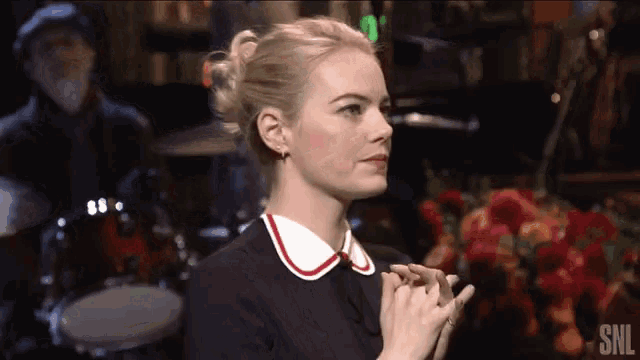 a woman in a black dress with a white collar is clapping her hands in front of a drum set .