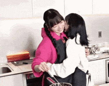 two girls are hugging in a kitchen and one is kissing the other on the cheek .