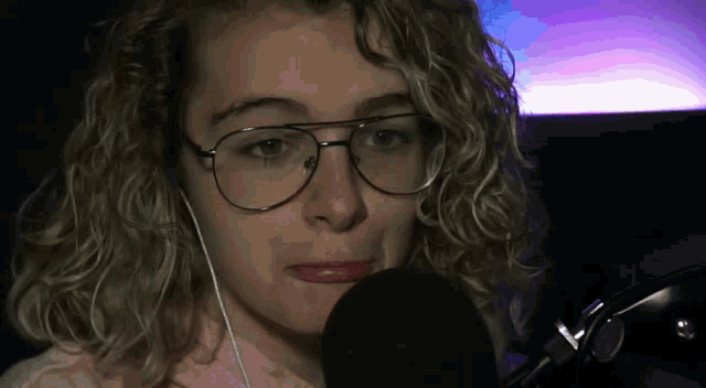 a woman with curly hair wearing glasses and ear buds talks into a microphone
