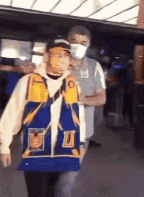 a man wearing a mask stands next to a man wearing a blue jacket with the letter u on it