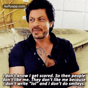 a man with a tattoo on his chest is sitting on a couch talking to someone .
