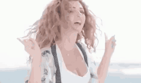 a woman with curly hair and a plunging neckline is standing on a beach with her hands in the air .