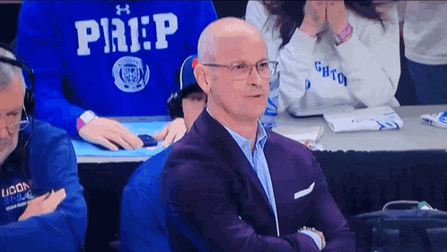 a man in a suit and glasses is sitting in front of a group of people wearing shirts that say prep
