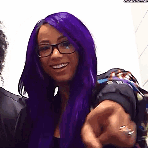 a woman with purple hair and glasses is pointing at the camera and smiling .