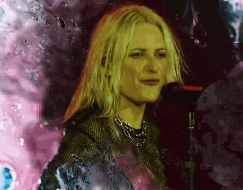 a blonde woman singing into a microphone with a pink background