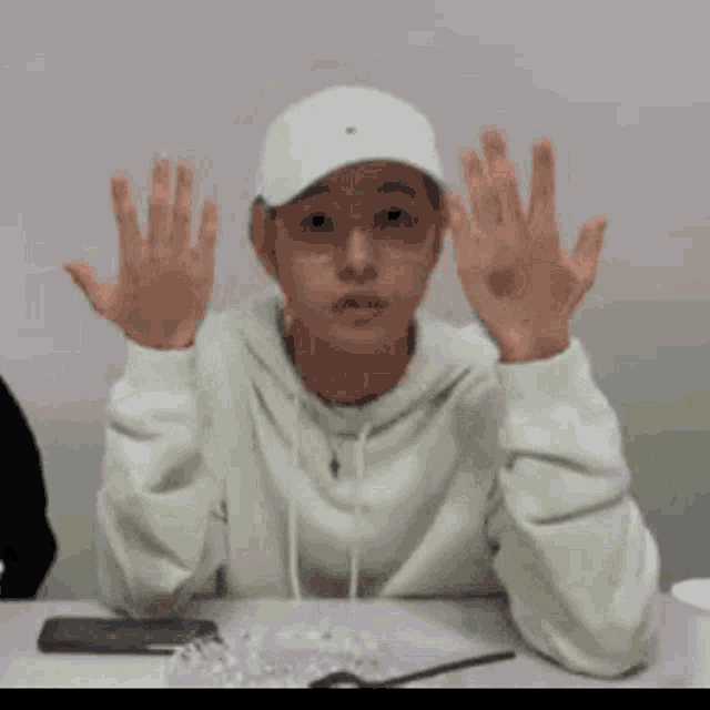 a young man wearing a white hat and glasses is sitting at a table with his hands up .