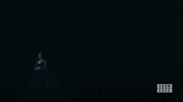 a woman is holding a light in her hand in a dark room with the show case logo in the corner .