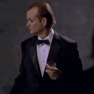a man in a tuxedo is holding a glass of whiskey while smoking a cigarette .
