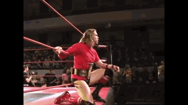 a man in a red shirt is kneeling in a wrestling ring holding a rope .