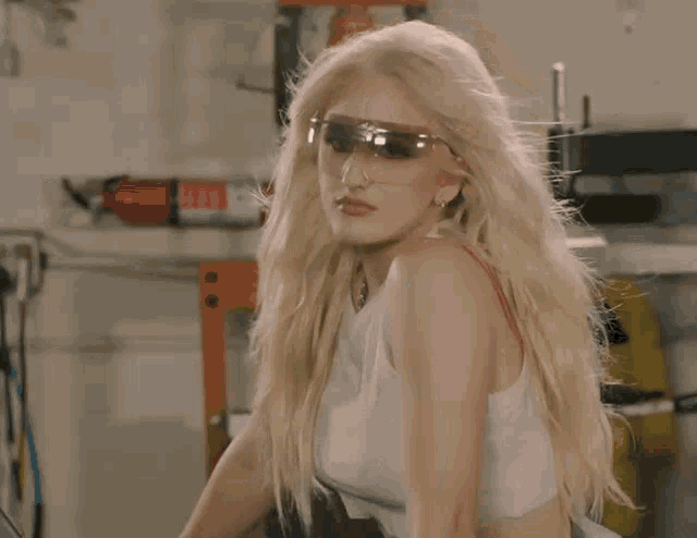 a blonde woman wearing a white tank top and safety goggles is standing in a garage .