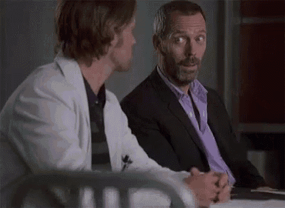two men are sitting at a table talking to each other . one of the men is wearing a purple shirt .
