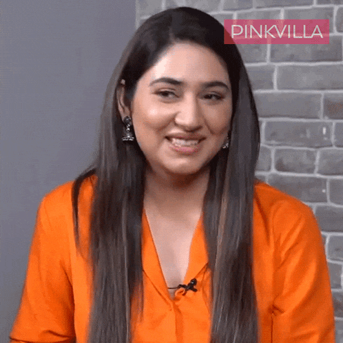 a woman wearing an orange shirt and earrings smiles in front of a brick wall with pinkvilla written on it