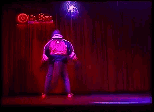 a man in a pink jacket is standing in front of a red curtain under a neon sign that says sol