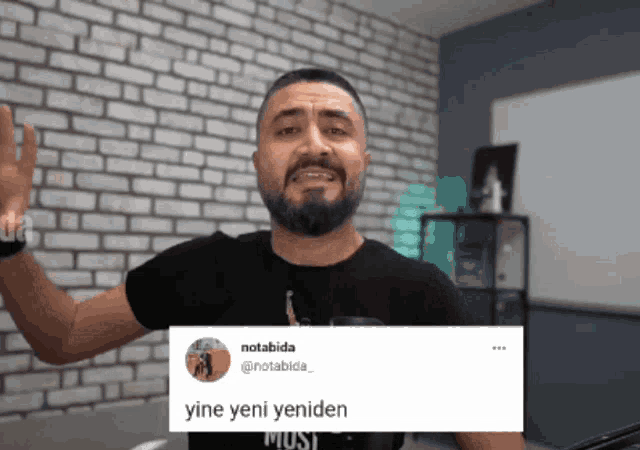 a man with a beard is standing in front of a brick wall holding a sign that says yine yeni yeniden
