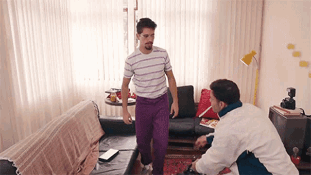 a man in purple pants is dancing in a living room with another man .