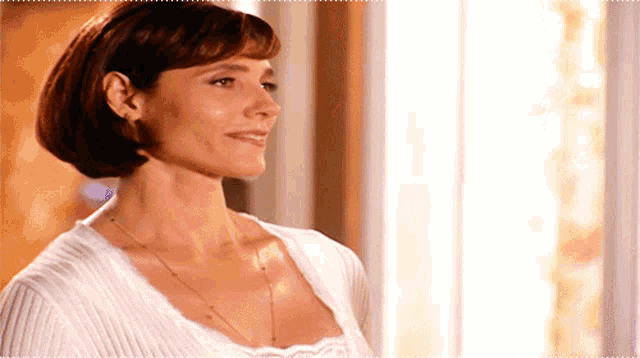 a woman wearing a white sweater and a necklace is standing in front of a window .