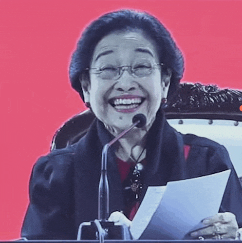 an older woman is smiling while holding a piece of paper and speaking into a microphone .