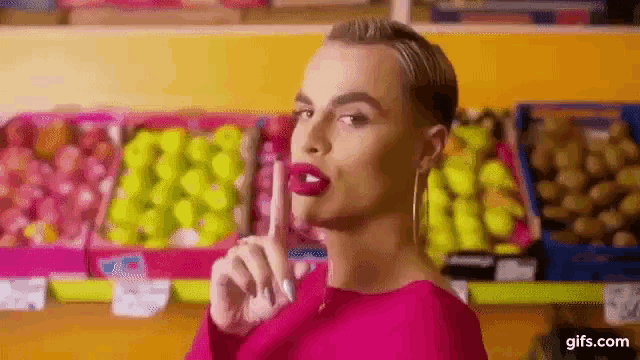 a woman in a pink dress is making a shhh gesture in front of a fruit stand .