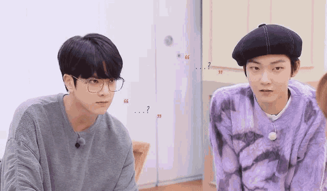 two young men wearing glasses and a beret are sitting next to each other and talking .
