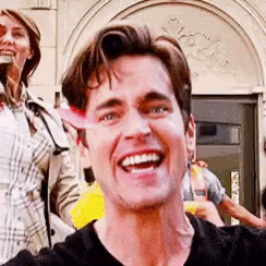 a man is smiling and taking a selfie in front of a building while a woman stands behind him .