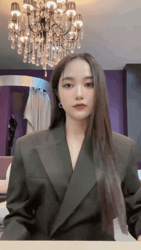 a woman in a suit stands in front of a chandelier in a room