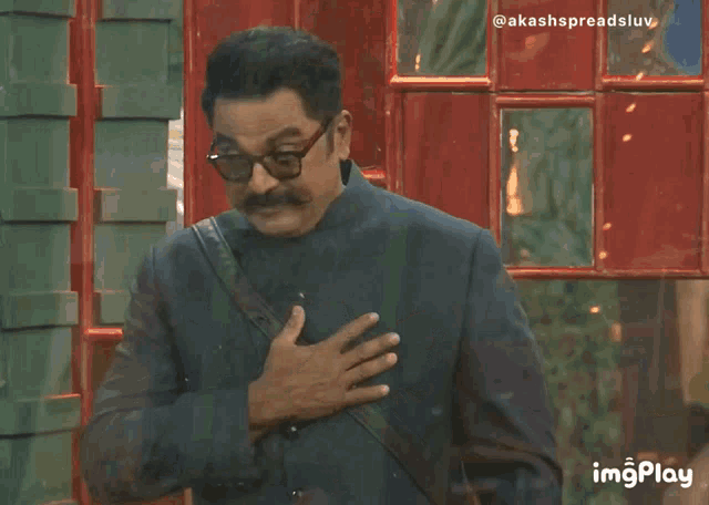 a man with glasses and a mustache holds his hands to his chest in front of a red phone booth