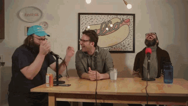 three men are sitting at a table with microphones and a picture of a hot dog on the wall behind them