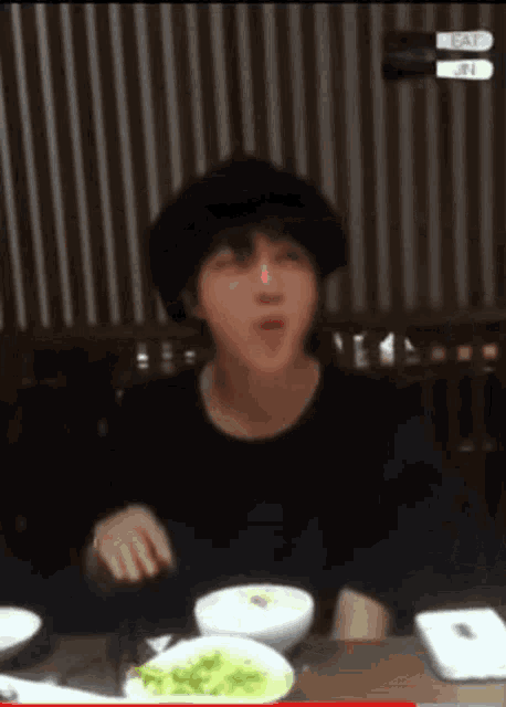 a man wearing a hat is sitting at a table with bowls of food and plates of food .