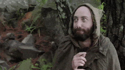 a man with a beard is wearing a poncho with a hood and standing in the woods .