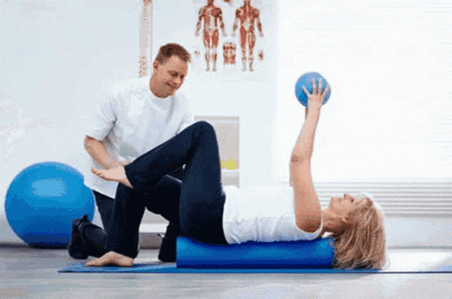 a woman is laying on a blue foam roller while a man holds a ball