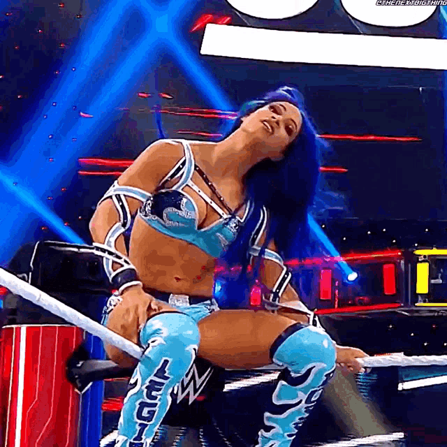 a female wrestler with blue hair is sitting on a rope in a wrestling ring .