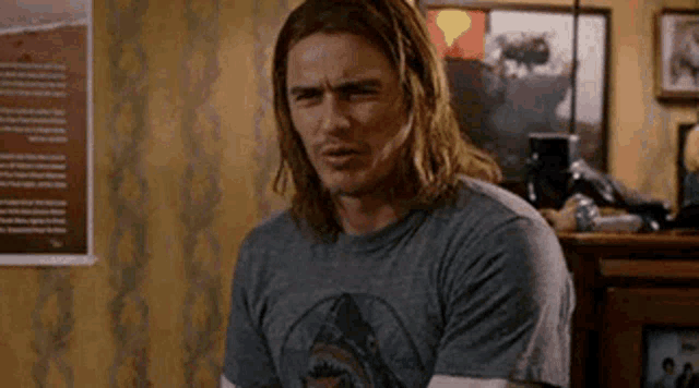 a man with long hair is wearing a shark t-shirt and sitting in a living room .