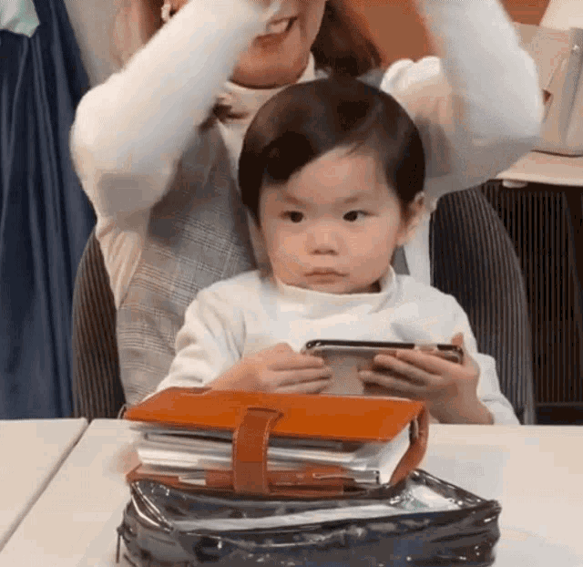 a little girl is sitting at a table holding a cell phone and a book .