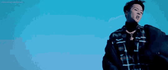 a young man is standing in front of a blue sky wearing a plaid shirt and a black jacket .