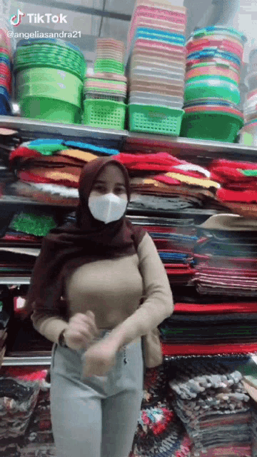 a woman wearing a hijab and a mask is standing in front of a shelf full of baskets and blankets .