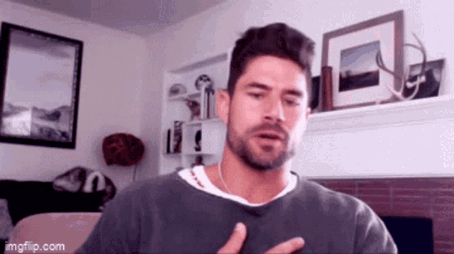 a man with a beard is in a living room with a fireplace and pictures on the wall .