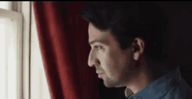 a man is looking out a window with a red curtain behind him .