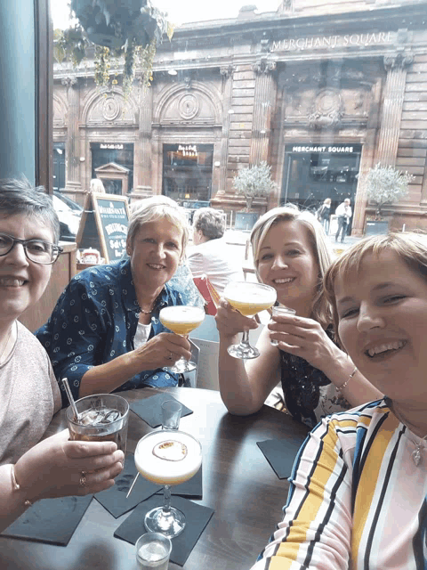 a group of women sitting at a table with drinks in front of a building that says merchant square