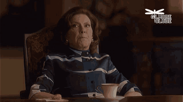 a woman is sitting at a table with a cup of coffee and a sign that says gilmore clube