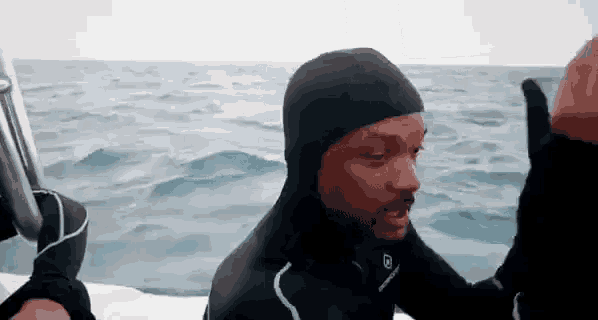 a man in a wetsuit is sitting on a boat in the ocean giving a thumbs up .